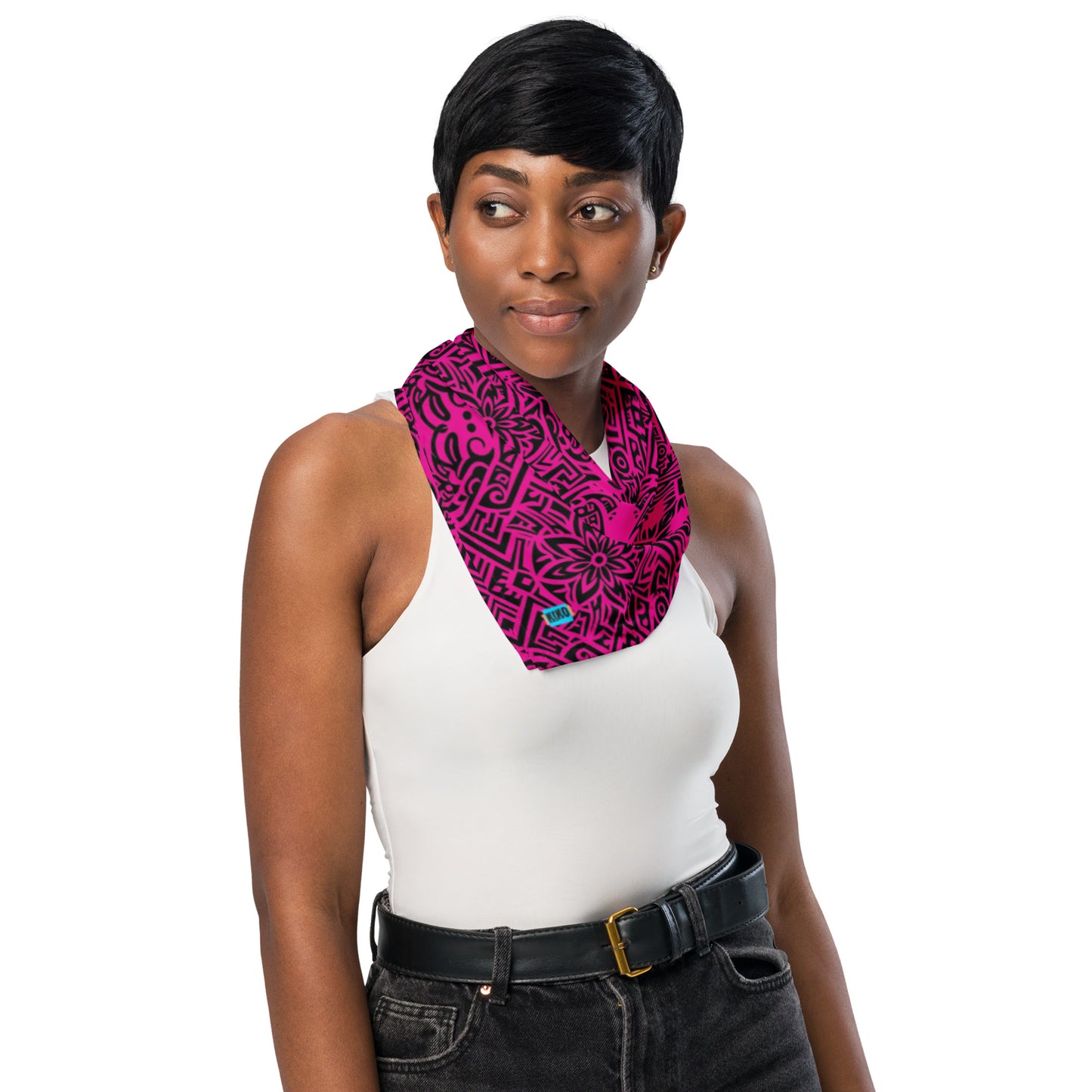 A young woman with short black hair stands against a plain white background, wearing a white sleeveless top, black pants with a black belt featuring a gold buckle, and a vibrant hot pink scarf bandana with intricate black hawaiian pattern around her neck.